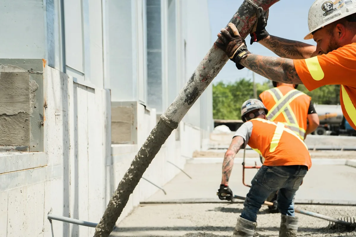 Workers in hard hats guiding a concrete pump hose while placing a slab beside a foundation wall